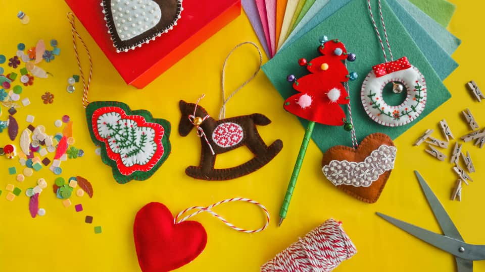 Several felt christmas decorations on a yellow table, in different shapes, including trees and hearts. There is felt, thread and scissors on the table around them.